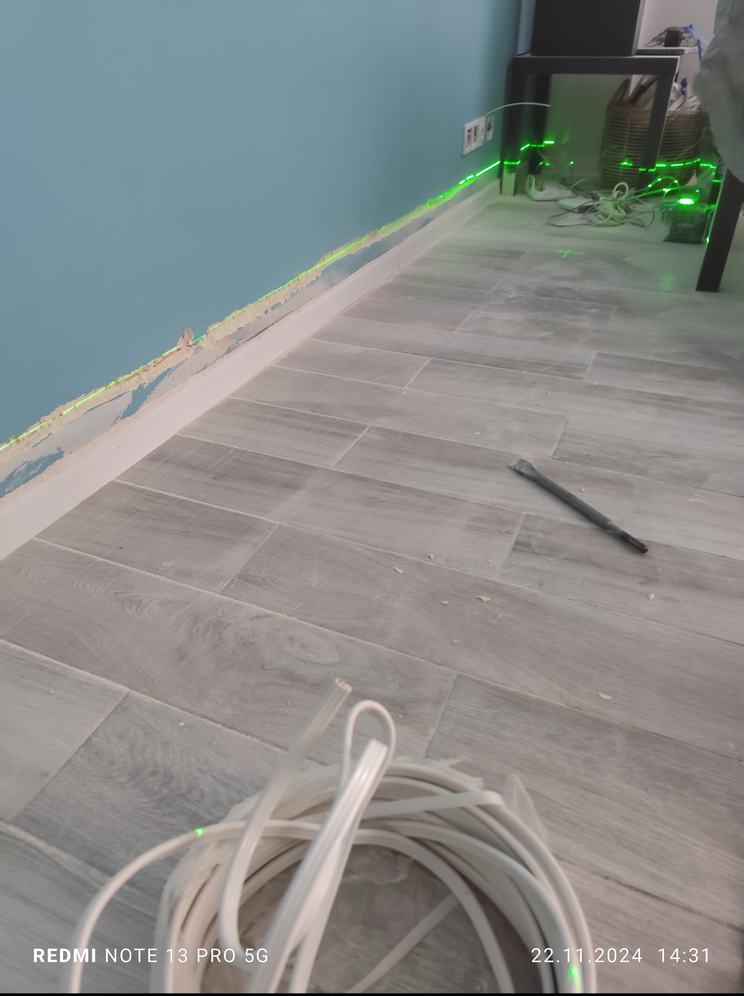 Przygotowanie do instalacji elektrycznej: wiązka kabli, dłuto i laserowy poziom na tle ściany z usuniętymi listwami przypodłogowymi i podłogi z płytek imitujących drewno.