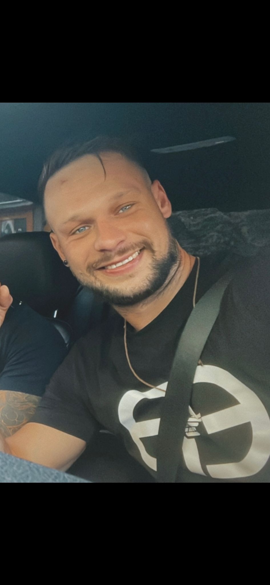 Uśmiechnięty mężczyzna z krótkimi włosami i lekkim zarostem, siedzący w samochodzie z zapiętym pasem bezpieczeństwa, ubrany w czarną koszulkę z białym nadrukiem.