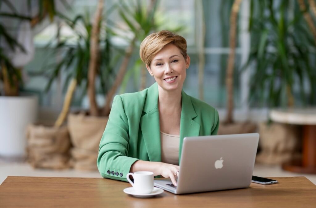 Uśmiechnięta blondynka w zielonej marynarce, siedząca przy biurku z laptopem i filiżanką kawy. W tle rośliny doniczkowe i okno.