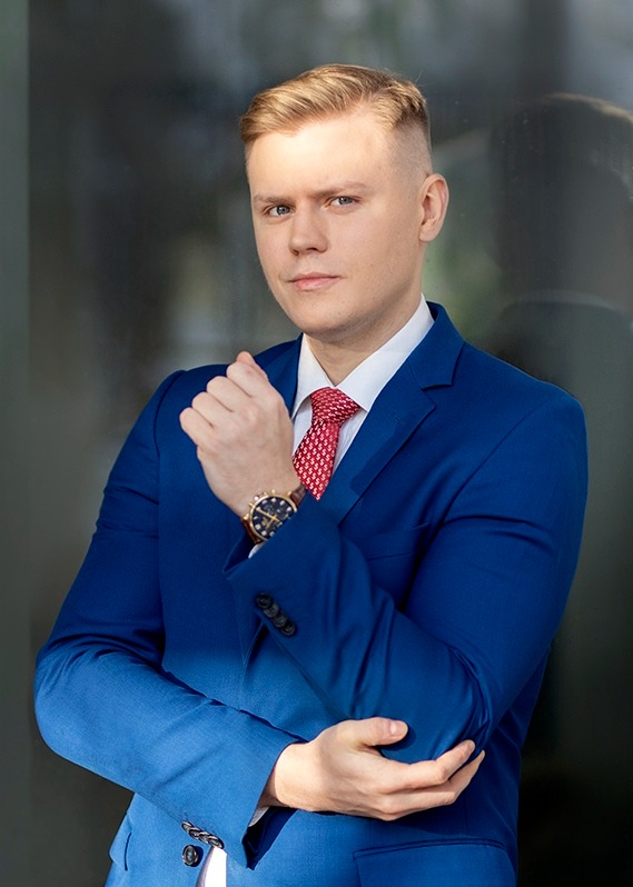 Mężczyzna w niebieskim garniturze i czerwonym krawacie, z zegarkiem na nadgarstku, pozuje na tle szklanej ściany. Profesjonalny wizerunek biznesowy.