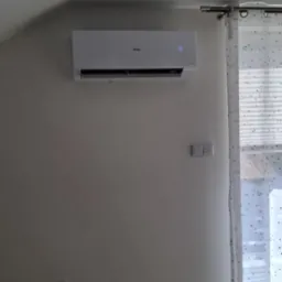 Biały klimatyzator Haier z wyświetlaczem 23°C, zamontowany na ścianie w jasnym pokoju, obok okna z zasłoną w gwiazdki. Widoczne gniazdka elektryczne.