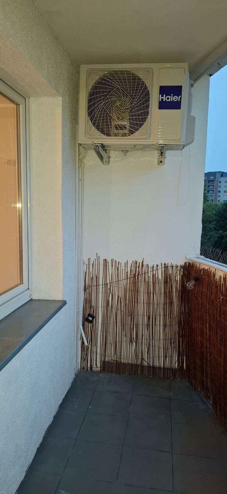 Biała klimatyzacja Haier zamontowana na balkonie z bambusową osłoną i szarymi płytkami podłogowymi, widok z dołu.
