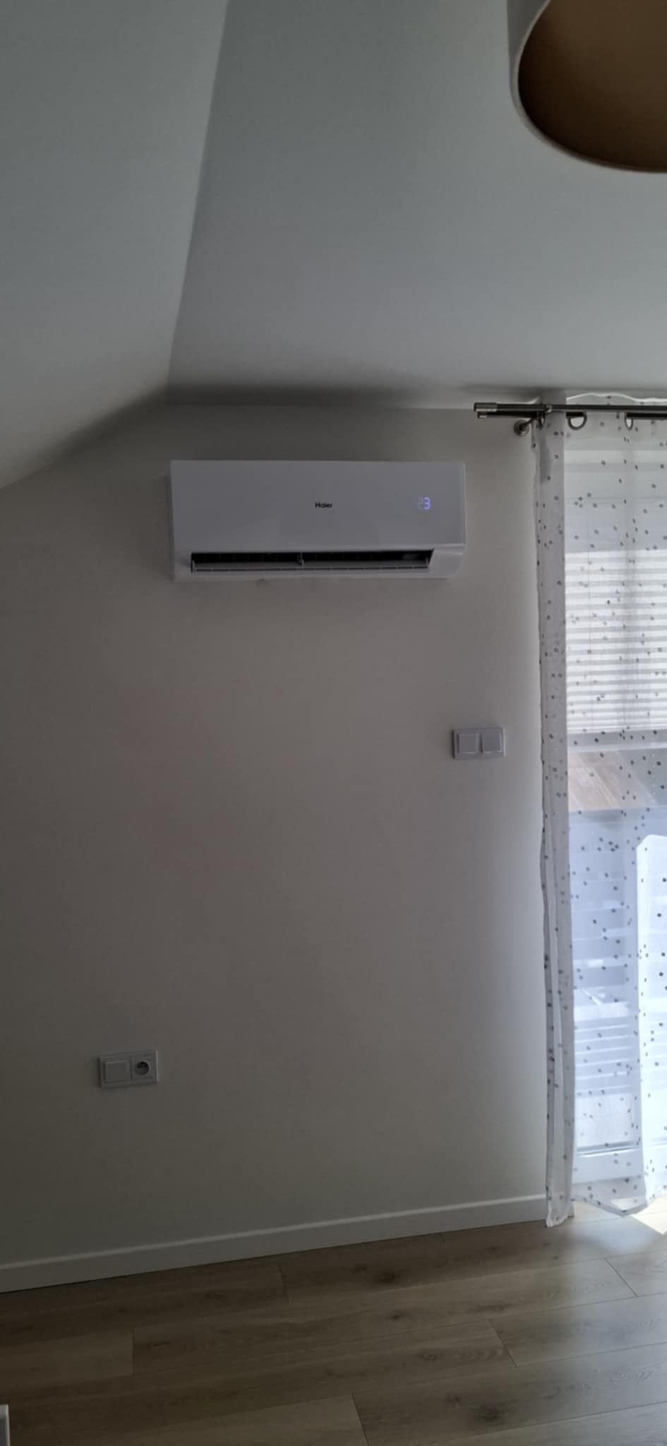 Biały klimatyzator Haier z wyświetlaczem 23°C, zamontowany na ścianie w jasnym pokoju, obok okna z zasłoną w gwiazdki. Widoczne gniazdka elektryczne.