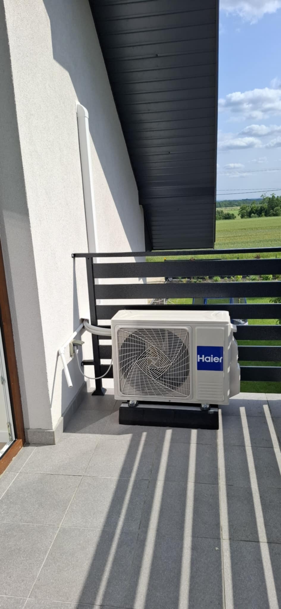 Biała jednostka zewnętrzna klimatyzacji Haier zamontowana na balkonie z widokiem na zielone pola. Widoczne przewody i cień balustrady.