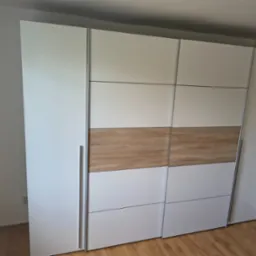 Nowoczesna szafa przesuwna z białymi frontami i drewnianym akcentem w środkowej części, z metalowymi uchwytami, na tle jasnej ściany i drewnianej podłogi.