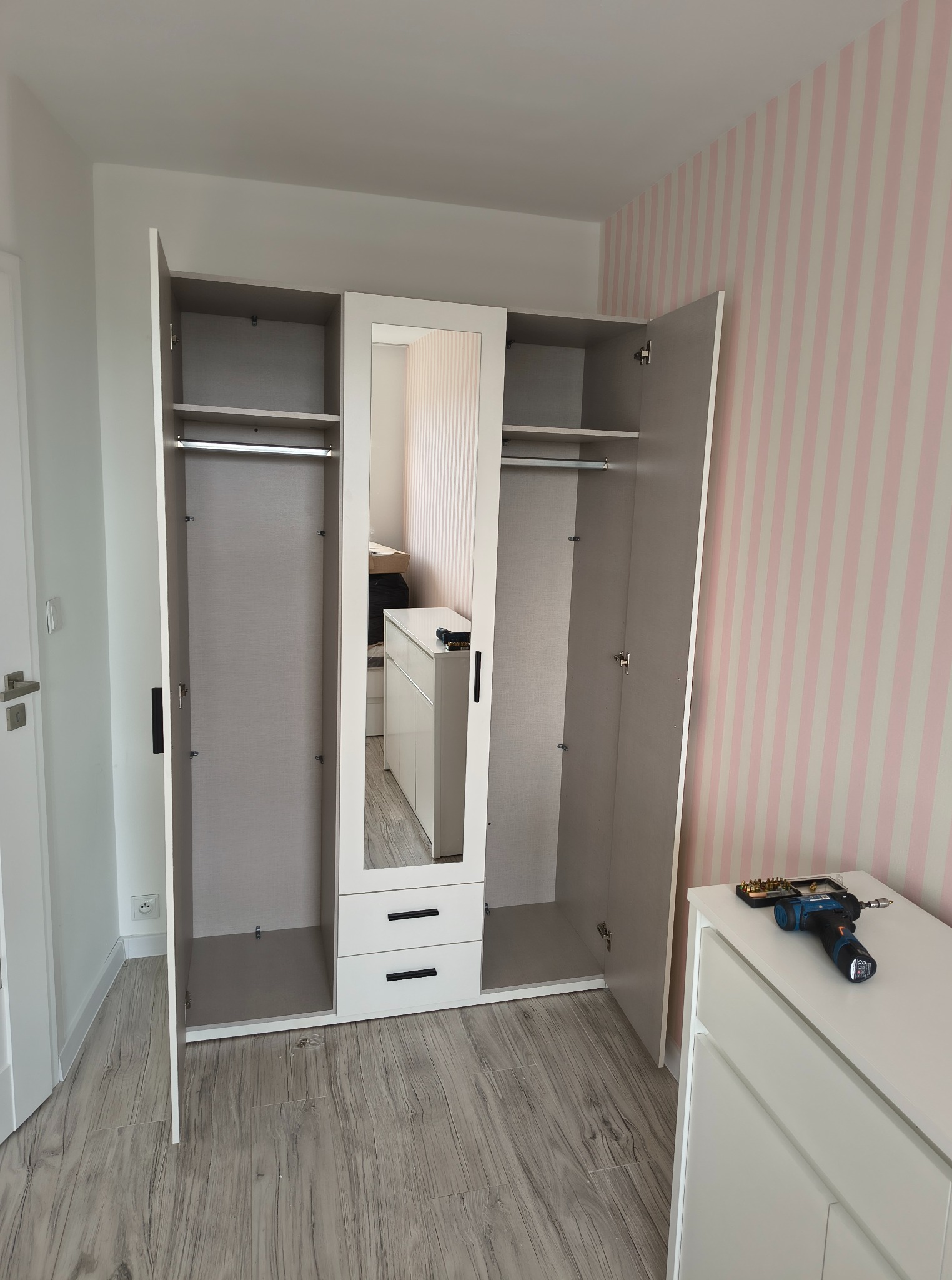 Nowoczesna, biało-szara szafa z lustrem i otwartymi drzwiami, na tle pokoju z różowo-białymi pasami. W tle widoczny wkrętak na komodzie.