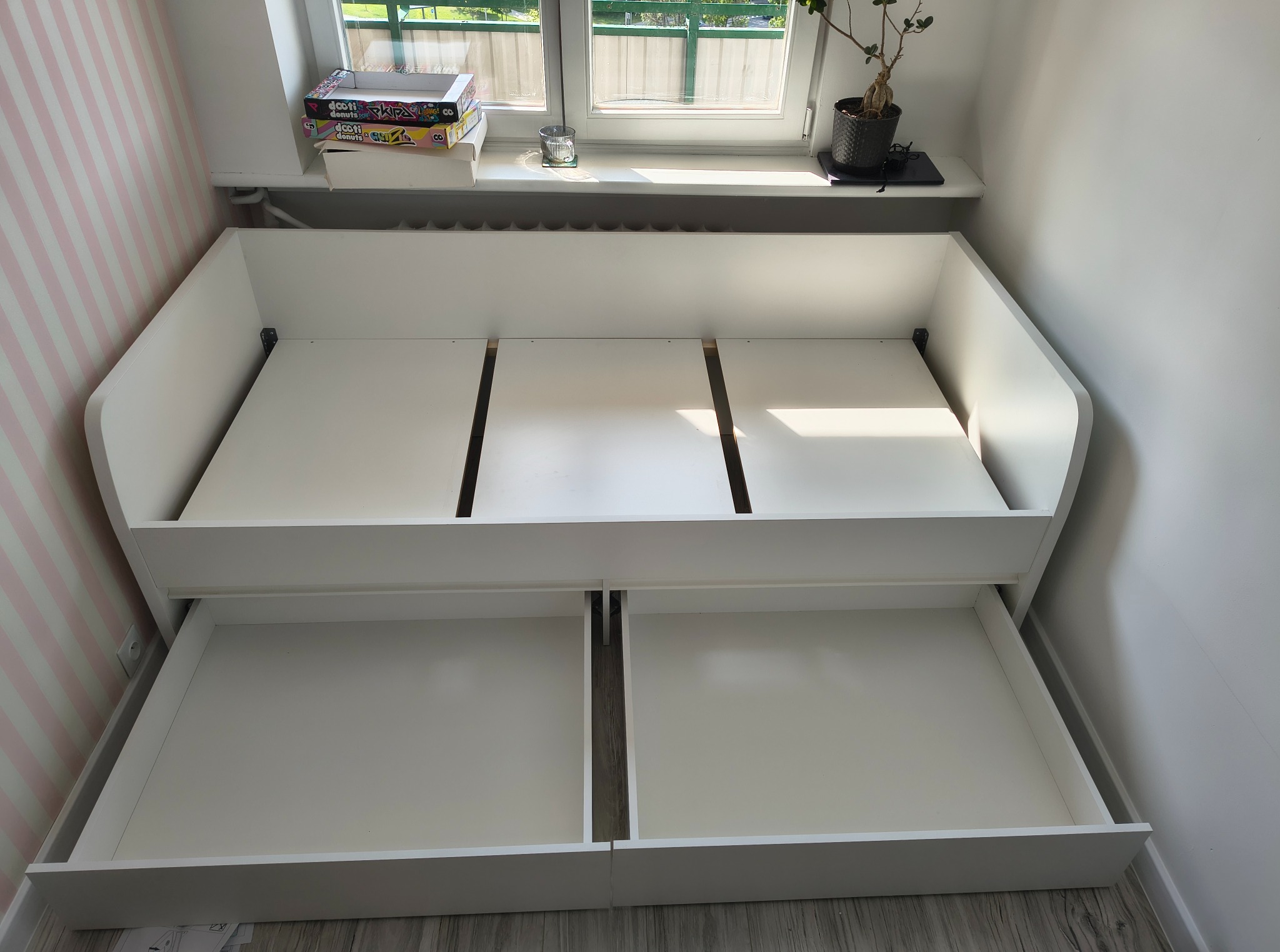 Białe, częściowo zmontowane łóżko z szufladami w pokoju z oknem, na tle ściany w różowe paski. Widoczny fragment podłogi.