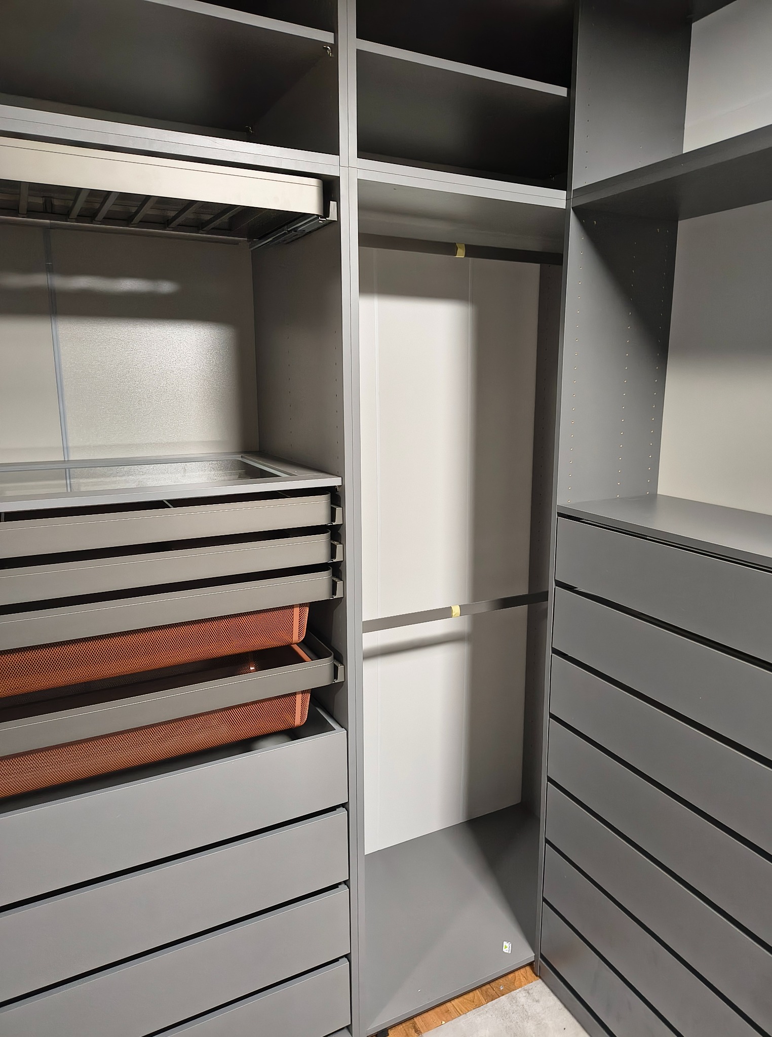 Szara, zabudowana garderoba z wysuwanymi półkami, szufladami i drążkami na ubrania. Widoczne detale wykończenia i praktyczne rozwiązania do przechowywania.