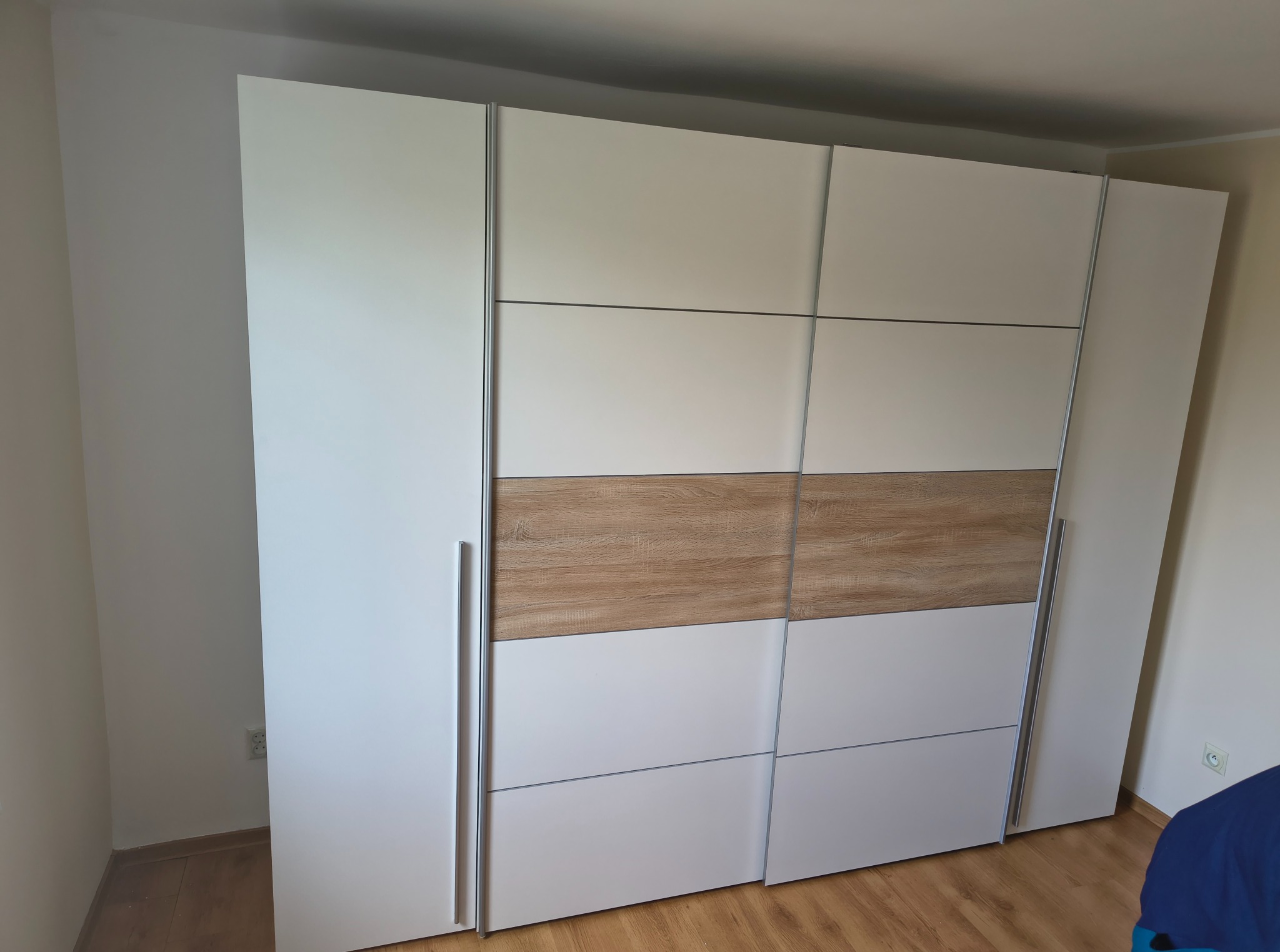 Nowoczesna szafa przesuwna z białymi frontami i drewnianym akcentem w środkowej części, z metalowymi uchwytami, na tle jasnej ściany i drewnianej podłogi.