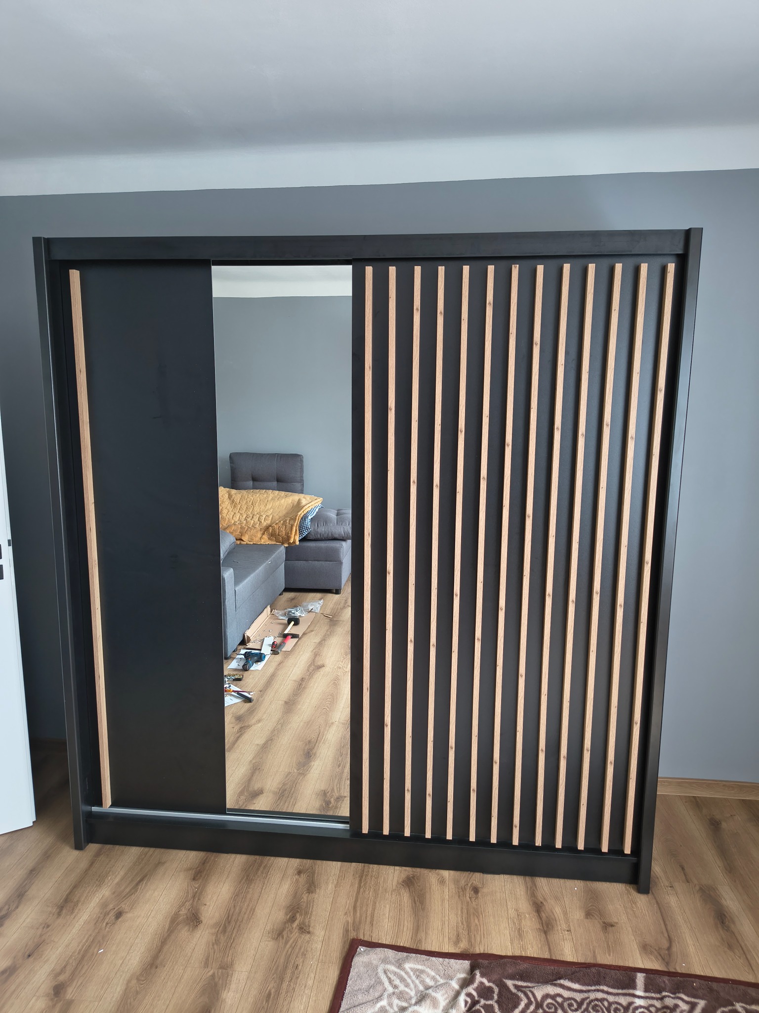 Czarna szafa przesuwna z lustrem i drewnianymi lamelami na tle szarej ściany. W lustrze odbicie pokoju z kanapą i narzędziami do montażu.