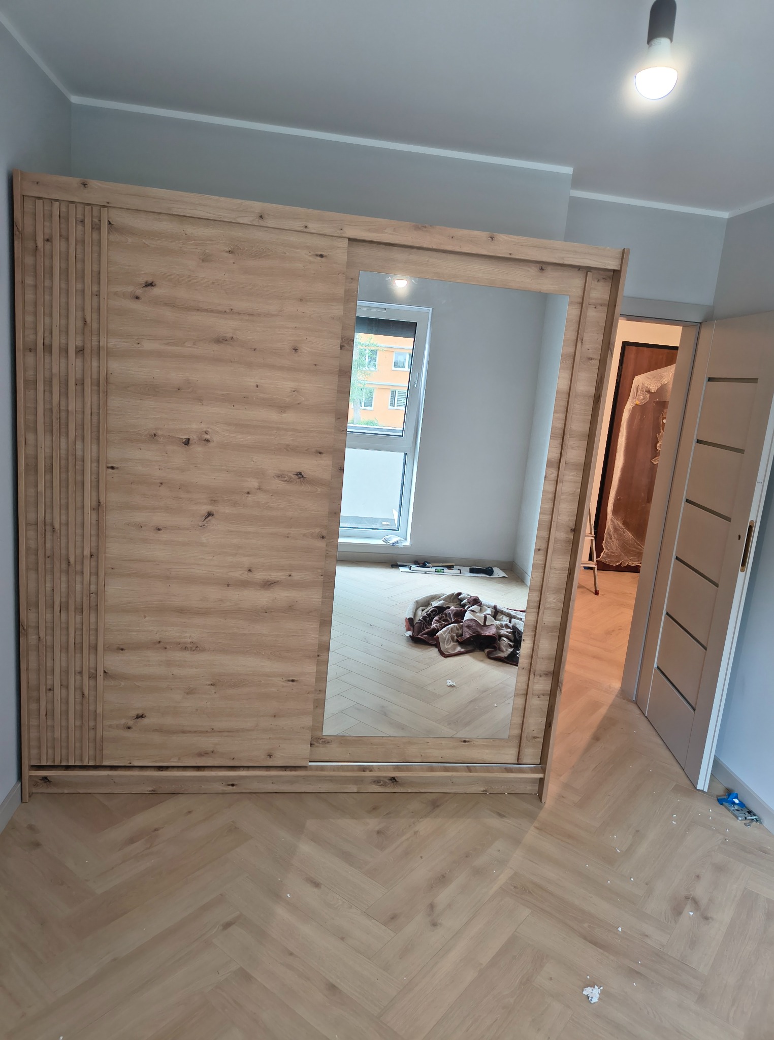 Jasna, drewniana szafa przesuwna z lustrem w pokoju z parkietem ułożonym w jodełkę. W tle widoczne okno i częściowo zasłonięte drzwi do innego pomieszczenia.