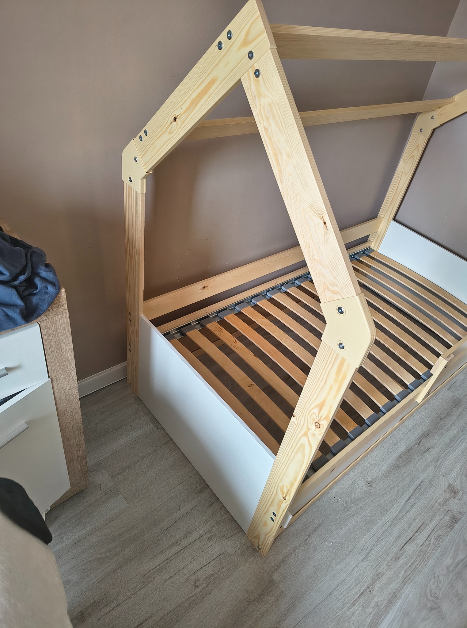 Drewniany, częściowo zmontowany stelaż łóżka w kształcie domku, z białym korpusem i drewnianymi listwami, stoi na podłodze z paneli, obok szafki nocnej.