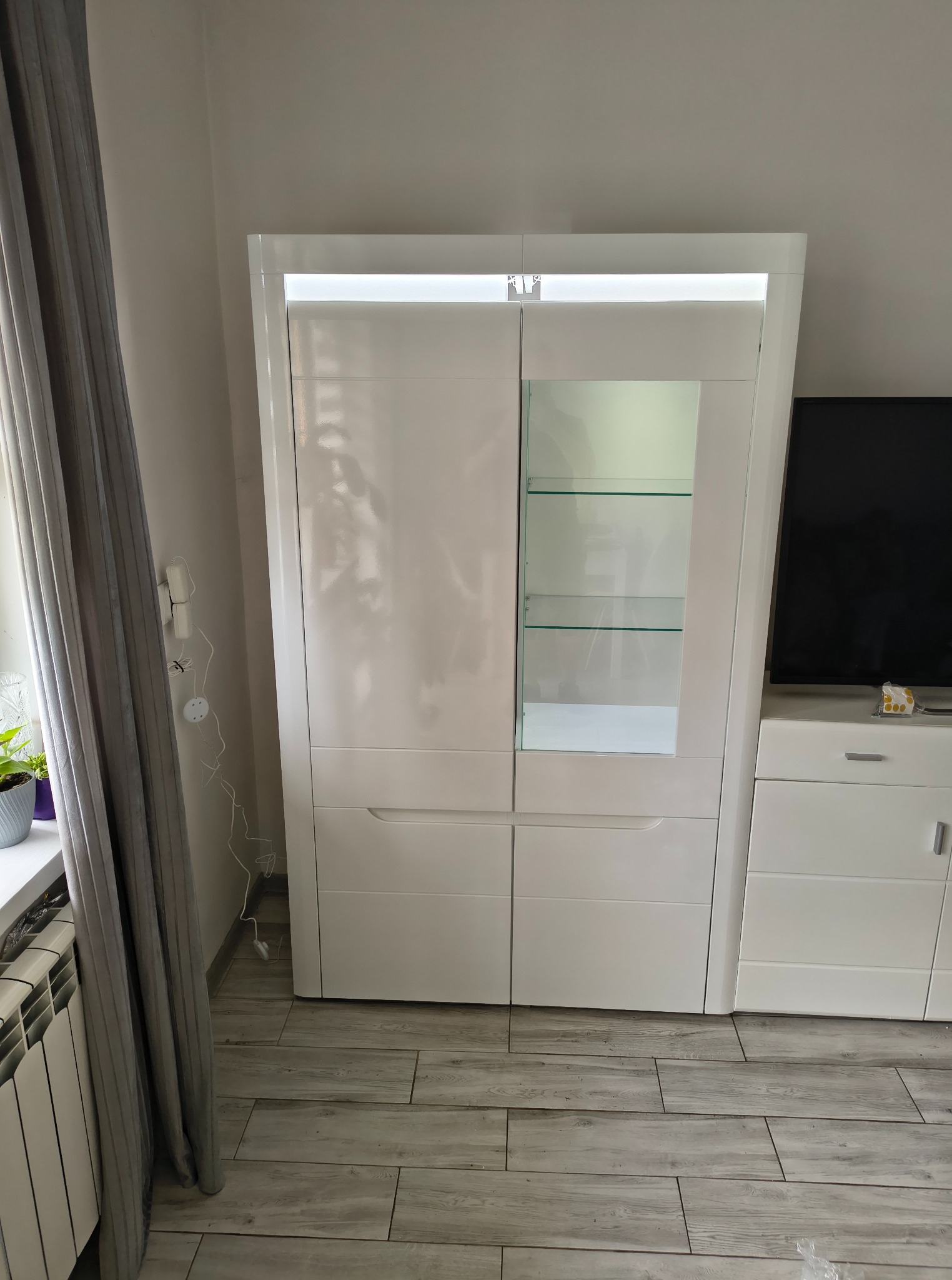 Biała, lakierowana witryna z oświetleniem LED w salonie. Widoczne szklane półki, szuflady i fragmenty innych mebli w jasnej tonacji. Ujęcie z bliska.