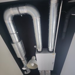 RefriTech - Widok z dołu na białą centralę wentylacyjną z podłączonymi, srebrnymi przewodami wentylacyjnymi na czarnym suficie, oświetloną przez dwie lampy.