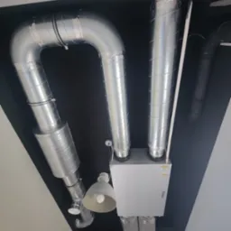 Widok z dołu na białą centralę wentylacyjną z podłączonymi, srebrnymi przewodami wentylacyjnymi na czarnym suficie, oświetloną przez dwie lampy.