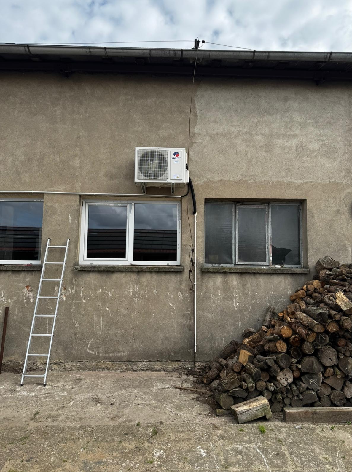Biały klimatyzator GREE na szarej ścianie budynku. Obok okno, drabina i stos drewna. Widoczne przewody instalacji. Pochmurne niebo w tle.