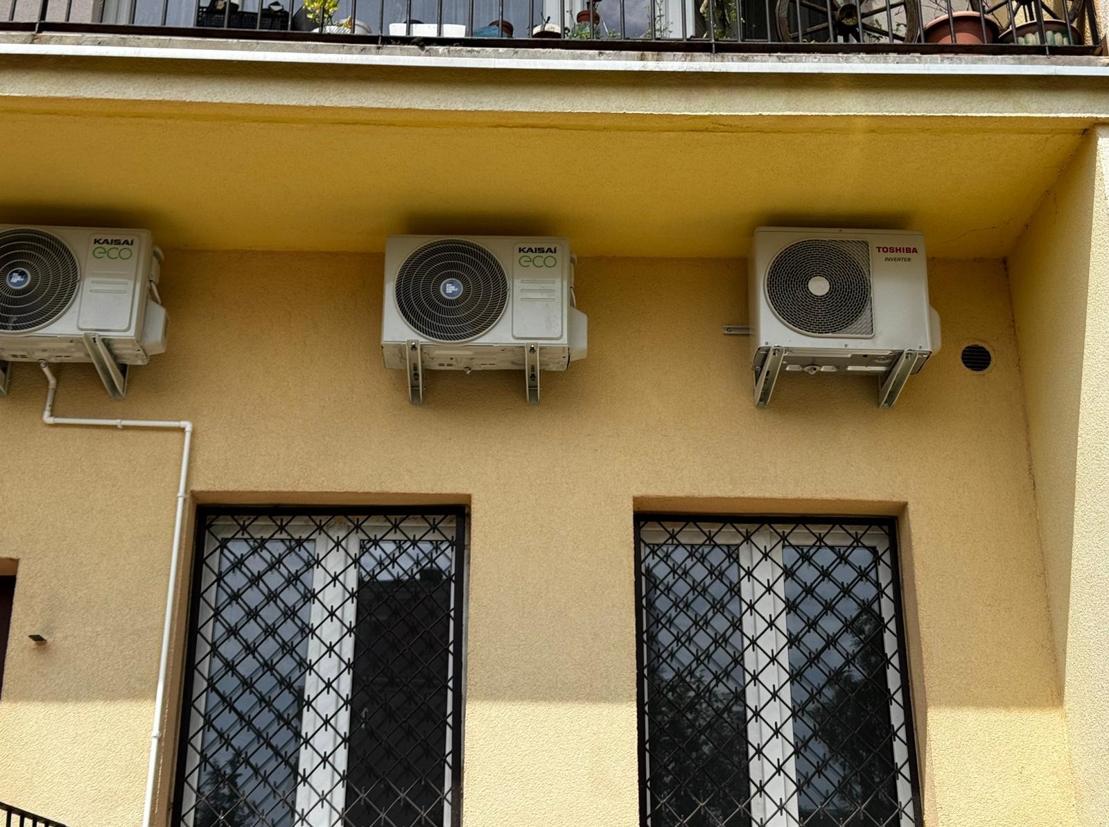 Elewacja budynku z trzema zamontowanymi klimatyzatorami (Kaisai Eco i Toshiba Inverter) nad oknami z kratami. Widoczny fragment balkonu z roślinami.