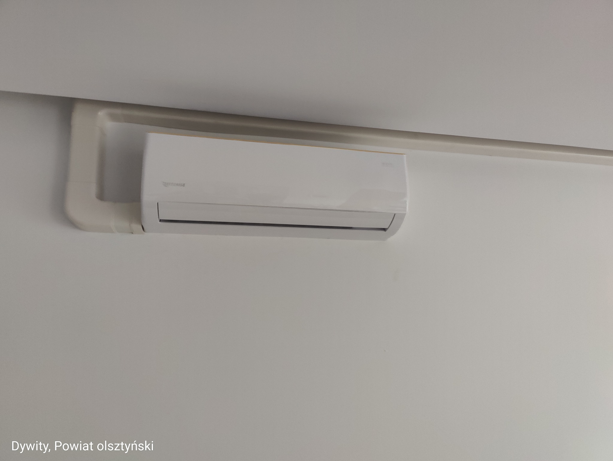 Biały klimatyzator na ścianie, widoczne rury instalacyjne. Całość w jasnej tonacji, z minimalnym oświetleniem, w tle biała ściana i fragment sufitu.
