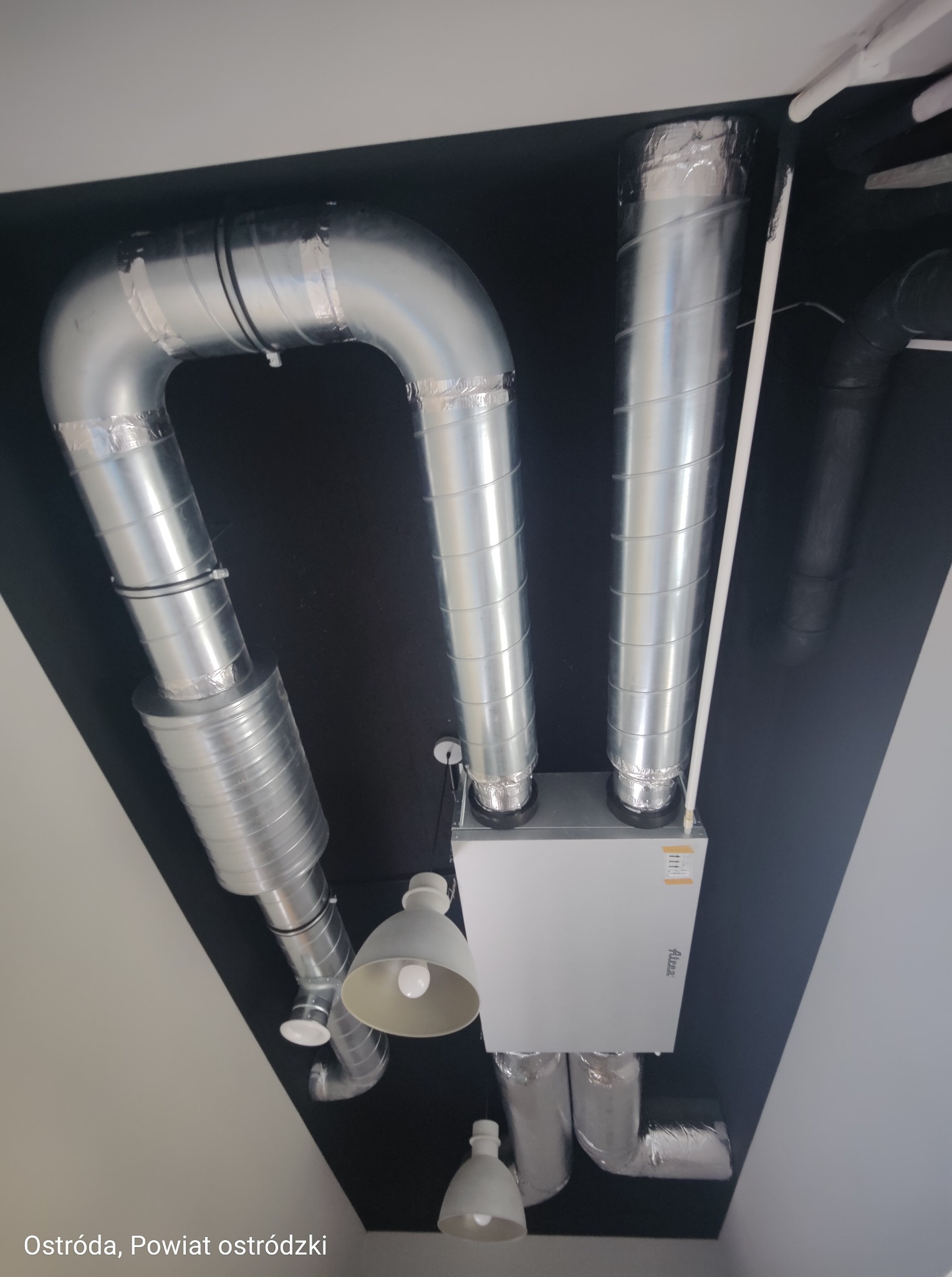 Widok z dołu na białą centralę wentylacyjną z podłączonymi, srebrnymi przewodami wentylacyjnymi na czarnym suficie, oświetloną przez dwie lampy.