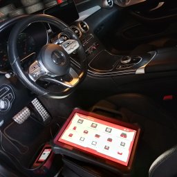 Q&L premium auto serwis - Wnętrze luksusowego auta, Mercedes-Benz. Na siedzeniu tablet diagnostyczny ThinkTool Euro Max z ikonami diagnostyki i serwisu. Widoczne pedały i kierownica.