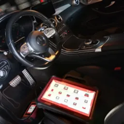 Wnętrze luksusowego auta, Mercedes-Benz. Na siedzeniu tablet diagnostyczny ThinkTool Euro Max z ikonami diagnostyki i serwisu. Widoczne pedały i kierownica.