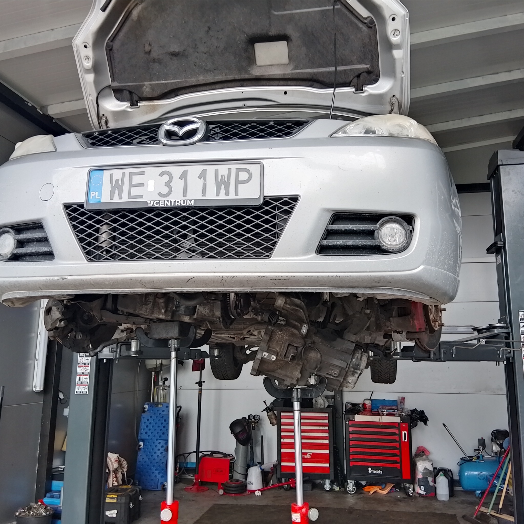 Srebrna Mazda podniesiona na podnośniku w warsztacie. Widoczne podwozie z elementami zawieszenia i skrzyni biegów. Otwarta maska. Tablica rejestracyjna WE 311WP.