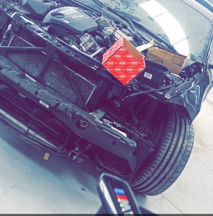 Czarny samochód z otwartą maską w warsztacie, widoczny silnik, chłodnica i części zamienne w pudełkach. Kąt ujęcia z dołu, oświetlenie warsztatowe.