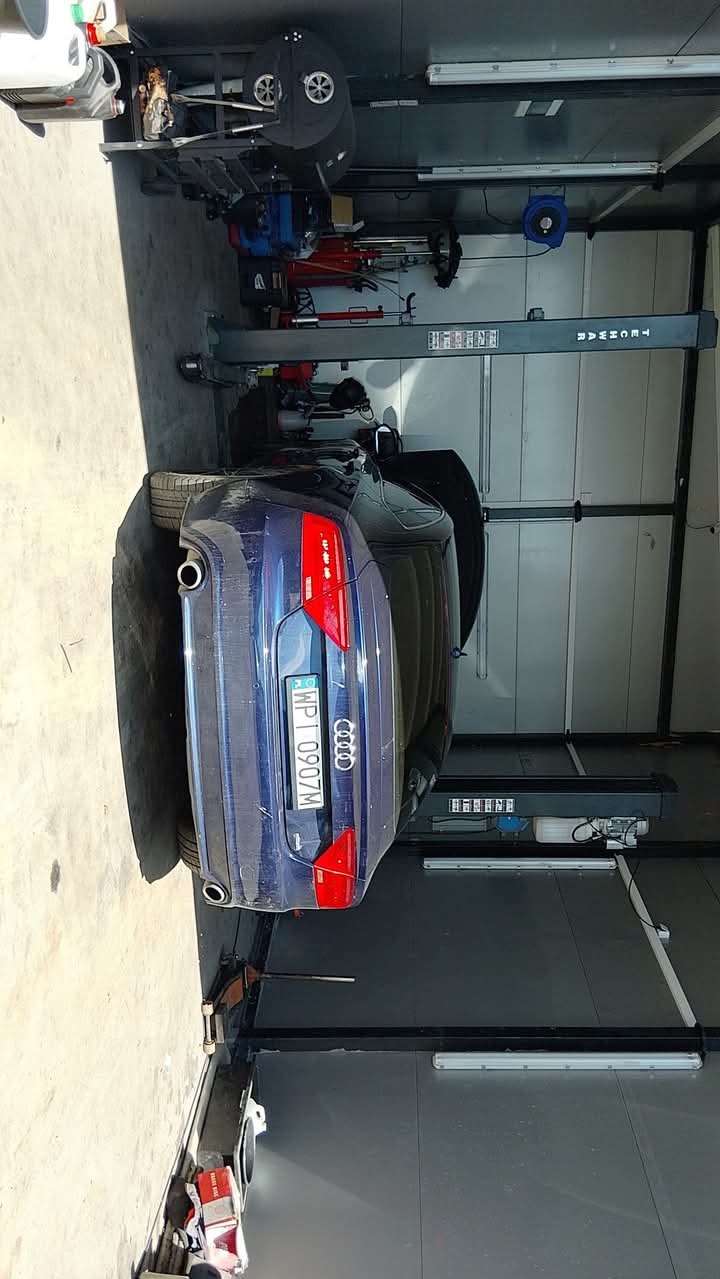 Niebieski samochód Audi z otwartą maską, widziany z tyłu, stoi na podnośniku w warsztacie. W tle narzędzia i wyposażenie warsztatowe.