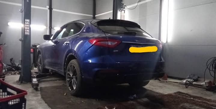 Niebieski samochód Maserati na podnośniku w warsztacie, widok z tyłu. Widoczne narzędzia i oświetlenie warsztatowe. Na podłodze ślady oleju.