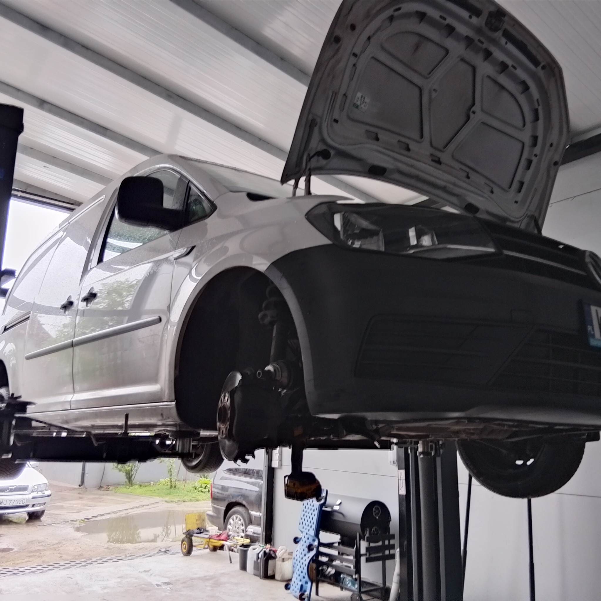 Samochód dostawczy na podnośniku w warsztacie, z otwartą maską i zdjętym kołem, ukazujący mechanizm zawieszenia. Widoczne narzędzia i wyposażenie warsztatu.