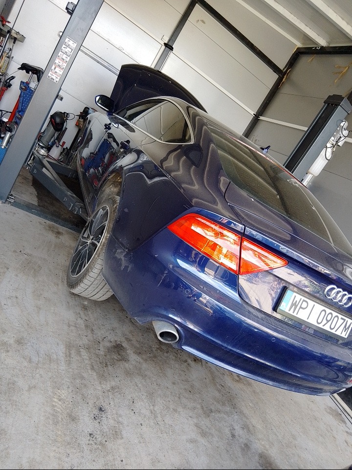 Niebieskie Audi A7 na podnośniku w warsztacie, z otwartą maską. Widoczny fragment lampy, koła i tablicy rejestracyjnej. Ujęcie z boku, lekko od tyłu.