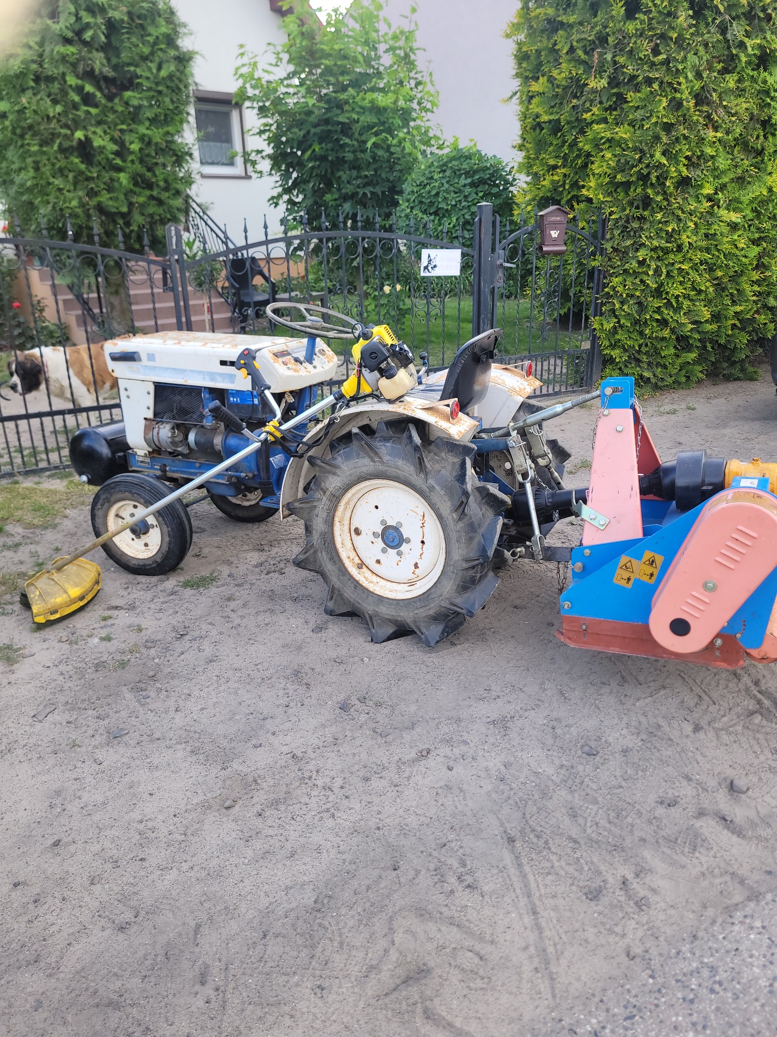 Mini traktorek z kosiarką listwową i glebogryzarką na szutrowej nawierzchni, widoczny również podkaszarka spalinowa i fragment psa za ogrodzeniem.