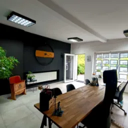 Nowoczesne biuro agencji nieruchomości z dużym drewnianym stołem, czarną ścianą z zegarem i zielonymi roślinami. W oknach widoczne oferty sprzedaży.