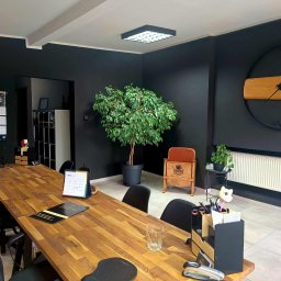 PENTHOUSE-Nieruchomości Katarzyna Bzura - Nowoczesne biuro nieruchomości z długim drewnianym stołem, czarnymi ścianami, zegarem i roślinami. Widoczny kalendarz i materiały firmowe.