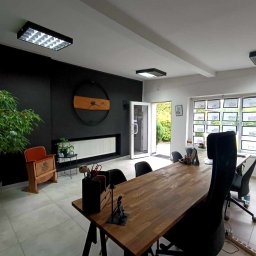 PENTHOUSE-Nieruchomości Katarzyna Bzura - Klimatyczne biuro: długa drewniana lada, krzesła biurowe, ściana z zegarem, rośliny doniczkowe i zdjęcia ofert nieruchomości w oknach. Jasne wnętrze.