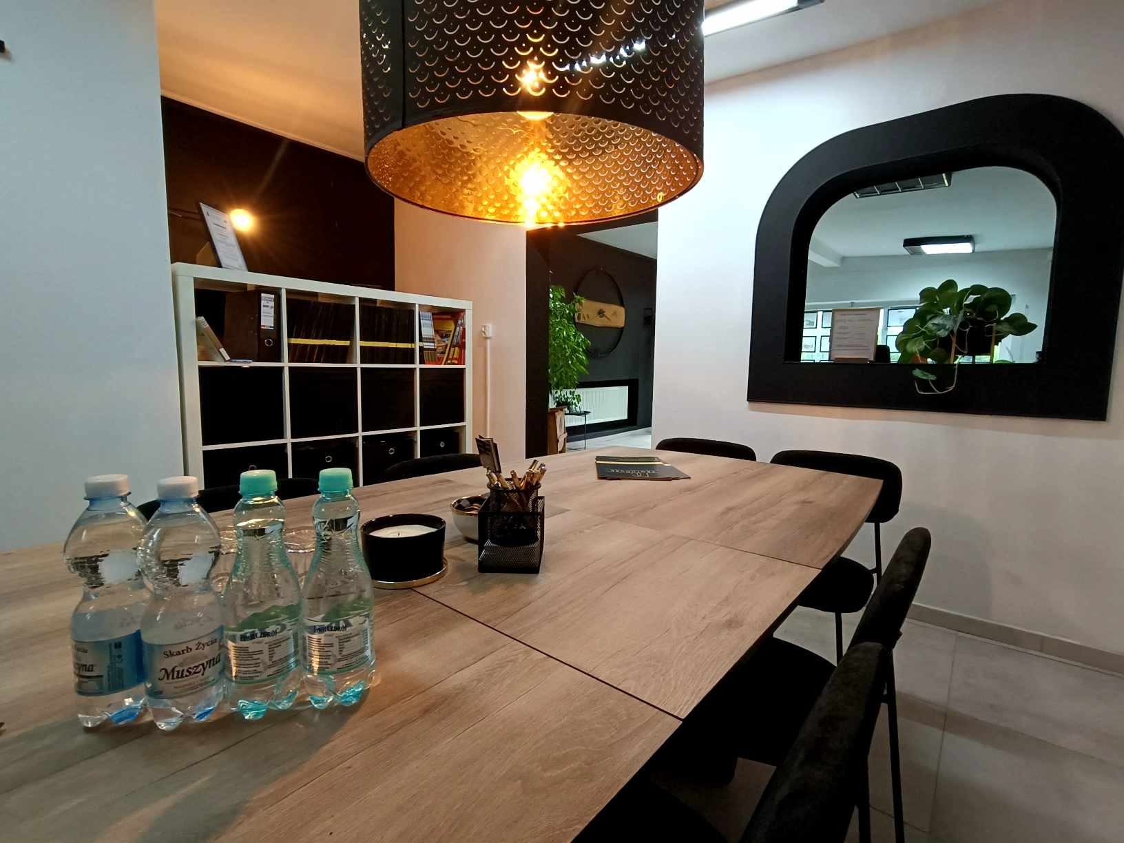 Sala konferencyjna z drewnianym stołem, czarnymi krzesłami i designerską lampą. Na stole woda i akcesoria biurowe. W tle regał i lustro w czarnej ramie.