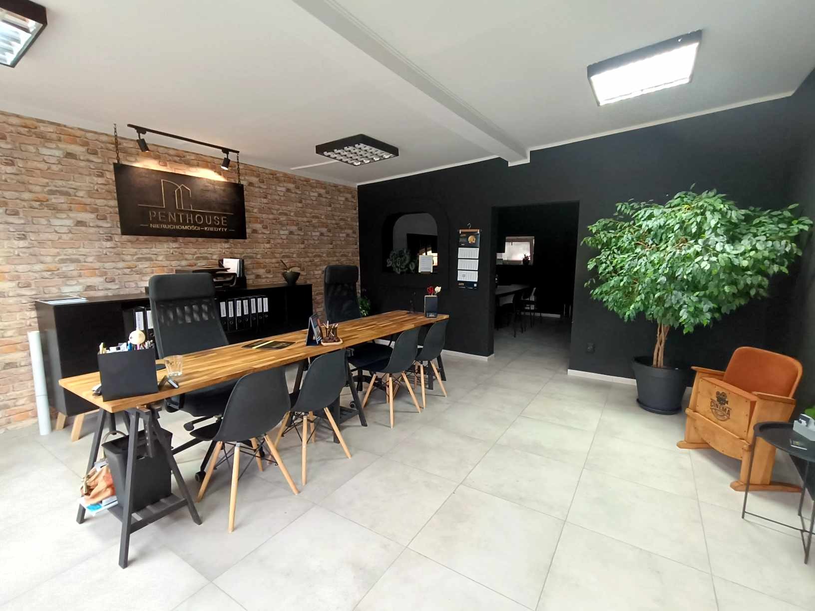 Nowoczesne biuro agencji nieruchomości z ceglaną ścianą, długim drewnianym stołem i czarnymi krzesłami. W tle widoczny napis 'Penthouse Nieruchomości-Kredyty'.