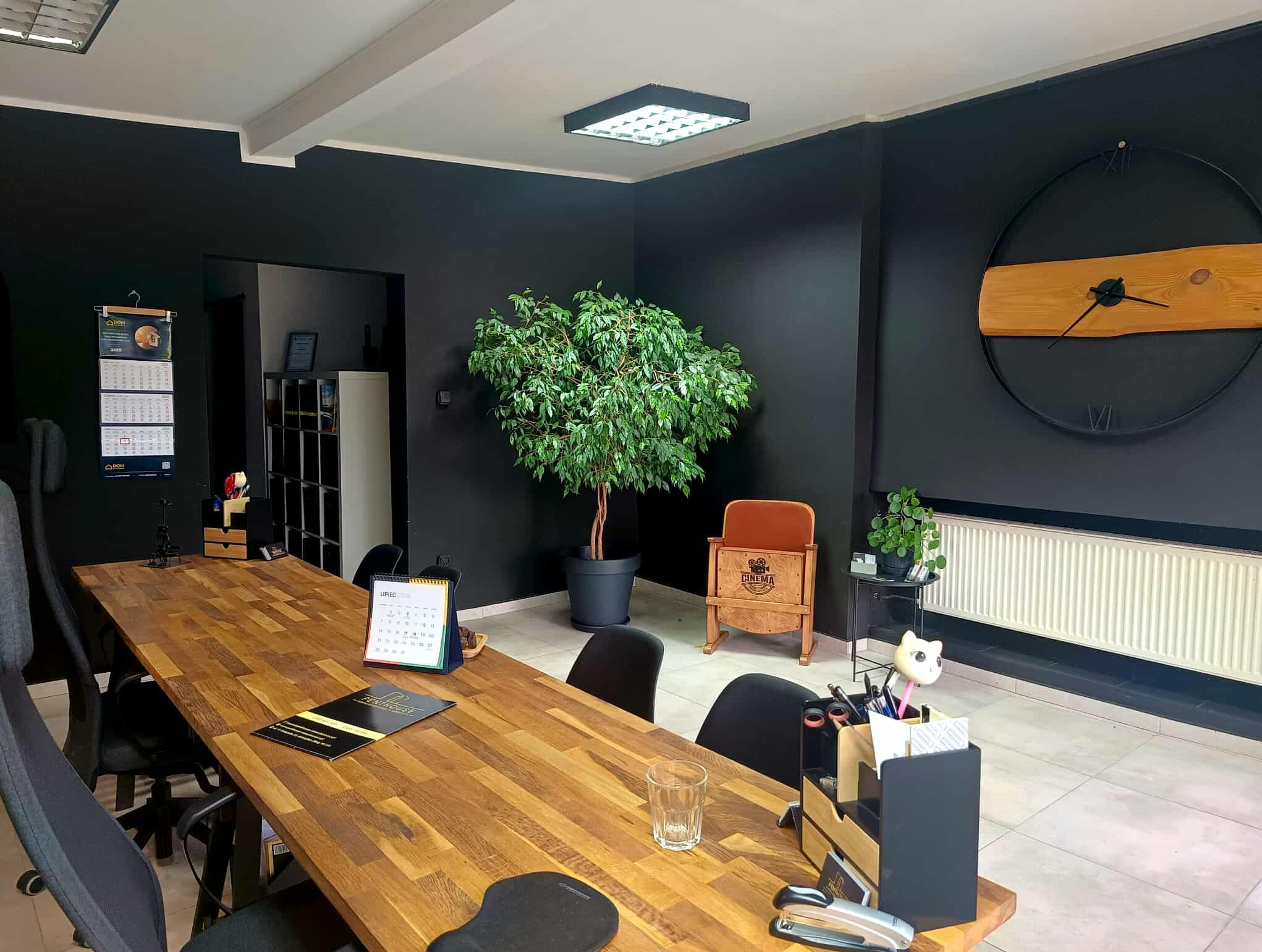 Nowoczesne biuro nieruchomości z długim drewnianym stołem, czarnymi ścianami, zegarem i roślinami. Widoczny kalendarz i materiały firmowe.