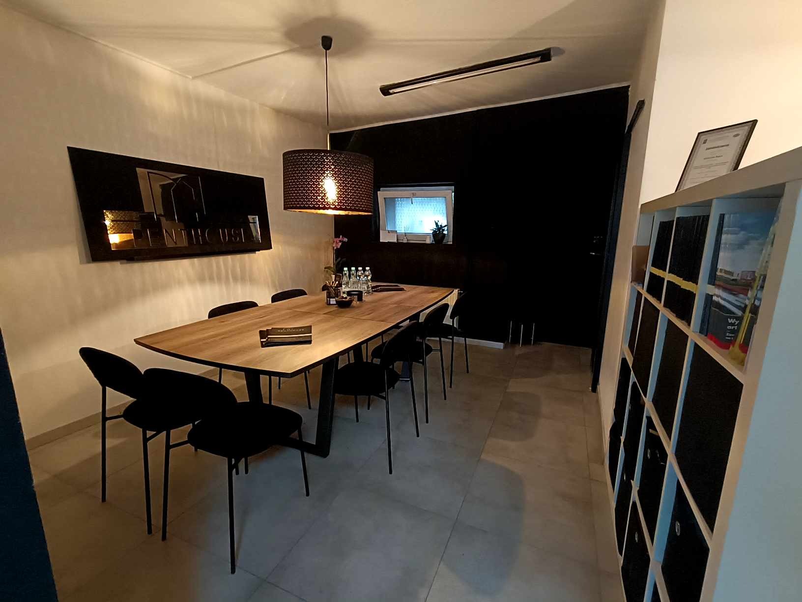 Nowoczesna sala konferencyjna z długim stołem z drewna i czarnymi krzesłami. Na ścianie dekoracja z nazwą firmy, oświetlenie punktowe i półki z dokumentami.