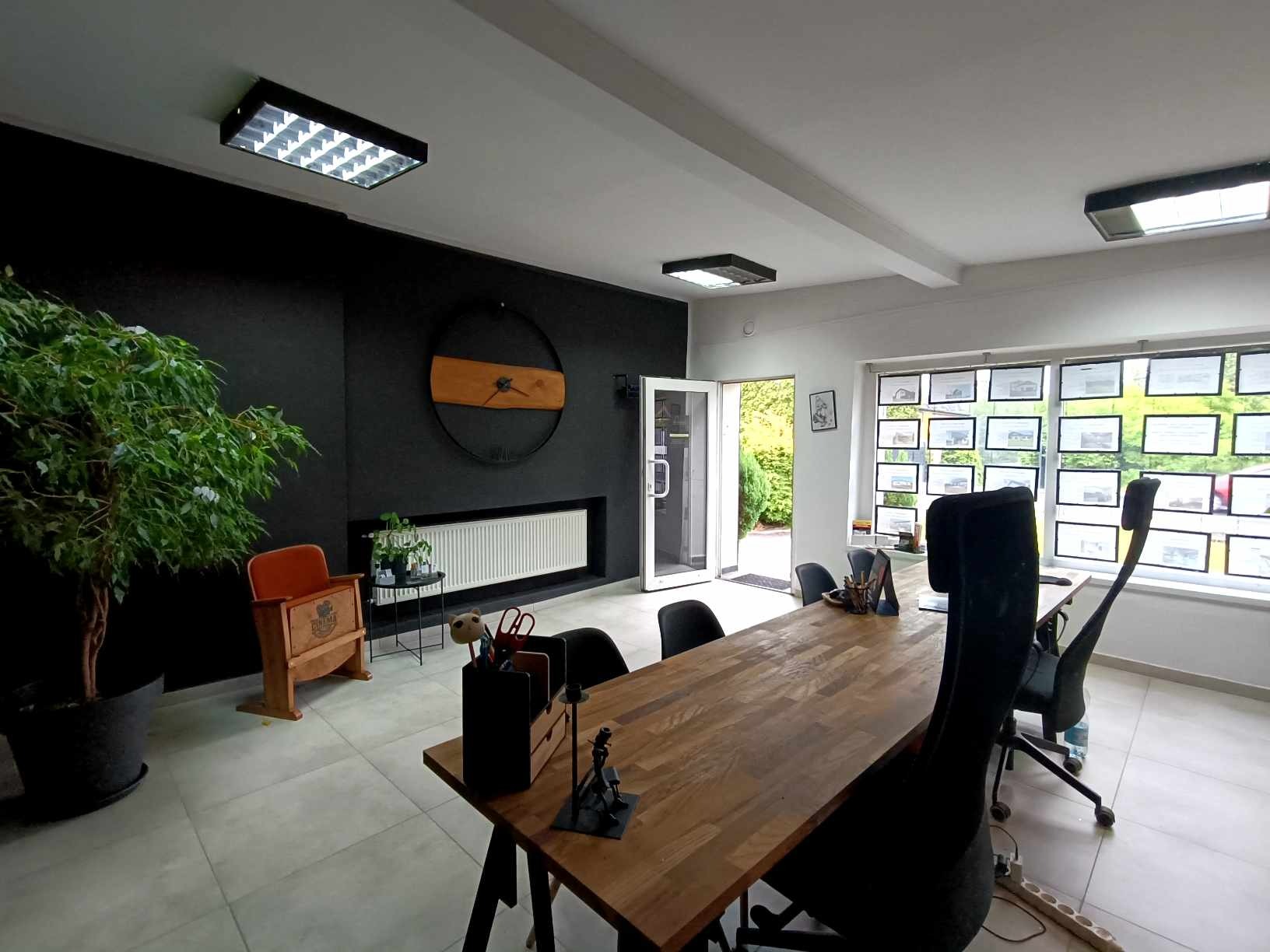 Klimatyczne biuro: długa drewniana lada, krzesła biurowe, ściana z zegarem, rośliny doniczkowe i zdjęcia ofert nieruchomości w oknach. Jasne wnętrze.