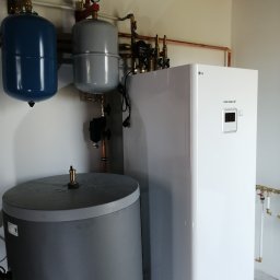 GREEN PLANET INSTALACJE - Biała pompa ciepła LG Therma V, szary zbiornik, niebieski i szary zbiornik wyrównawczy z manometrami. Miedziane rury instalacji grzewczej na ścianie.