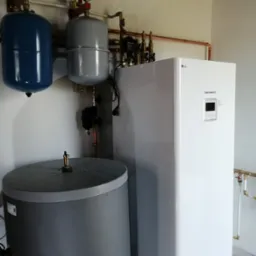 Biała pompa ciepła LG Therma V, szary zbiornik, niebieski i szary zbiornik wyrównawczy z manometrami. Miedziane rury instalacji grzewczej na ścianie.