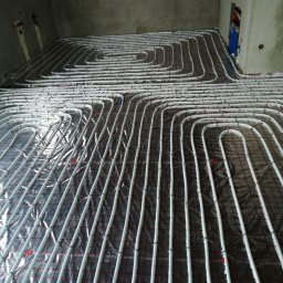 GREEN PLANET INSTALACJE - Białe rury ogrzewania podłogowego ułożone na folii z czerwoną kratą w surowym wnętrzu. Widoczne mocowania rur i fragment instalacji na ścianie.