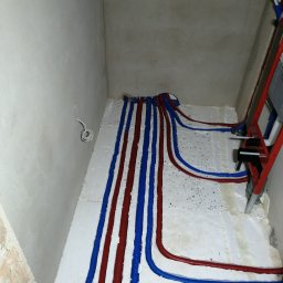 GREEN PLANET INSTALACJE - Montaż instalacji ogrzewania podłogowego z rurami w kolorach niebieskim i czerwonym, ułożonymi na białej izolacji, widok z góry w surowym wnętrzu łazienki.
