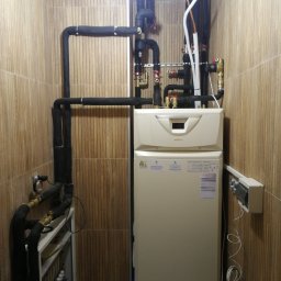 GREEN PLANET INSTALACJE - Zamknięte pomieszczenie z zamontowaną pompą ciepła Immergas, rurami w izolacji i rozdzielaczem ogrzewania podłogowego na tle ściany wyłożonej płytkami.