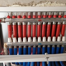 GREEN PLANET INSTALACJE - Rozdzielacz ogrzewania podłogowego z czerwonymi i niebieskimi rurami w metalowej szafce, widoczne zawory i mierniki przepływu. Instalacja w surowym wnętrzu.