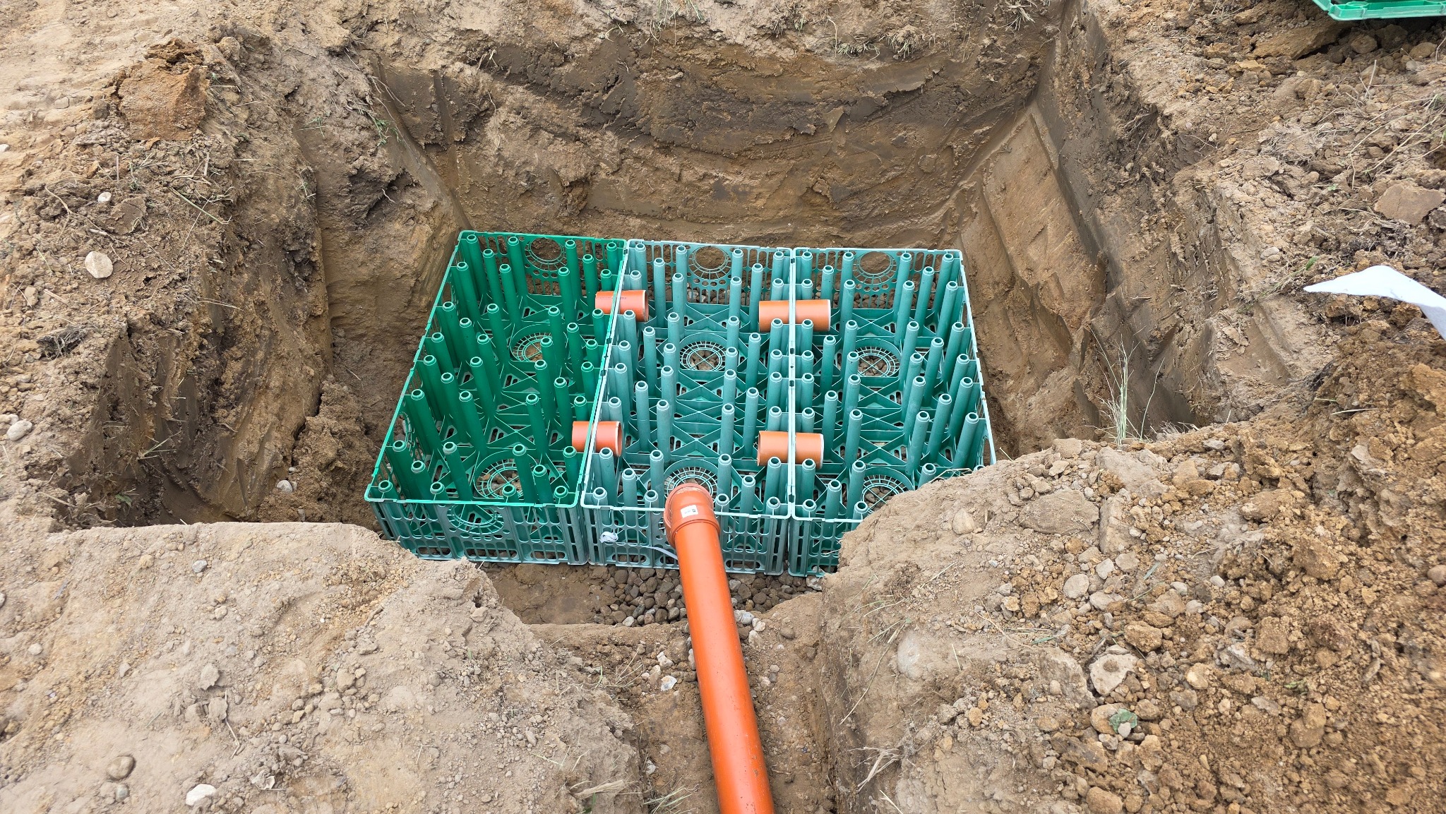 Instalacja drenażowa: zielone moduły retencyjne w wykopie, połączone pomarańczowymi rurami. Widok z góry na system drenażowy w ziemi.