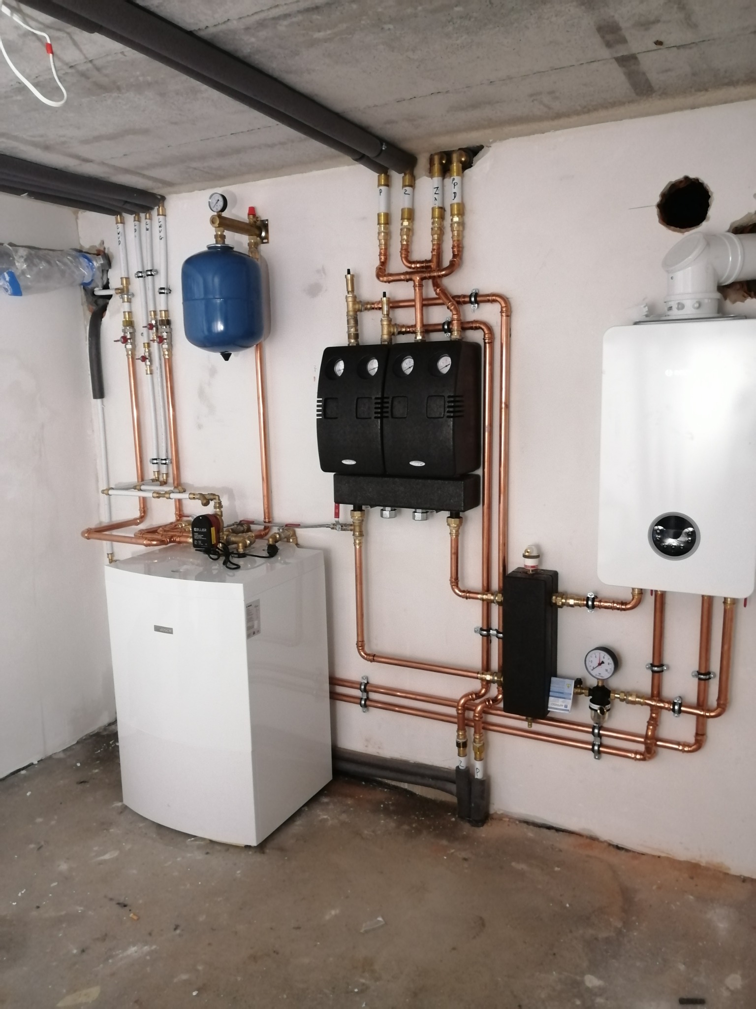 Kompletna instalacja grzewcza z piecem, zasobnikiem i rurami miedzianymi, widoczne manometry i zawory. Surowy betonowy strop i ściany w tle.