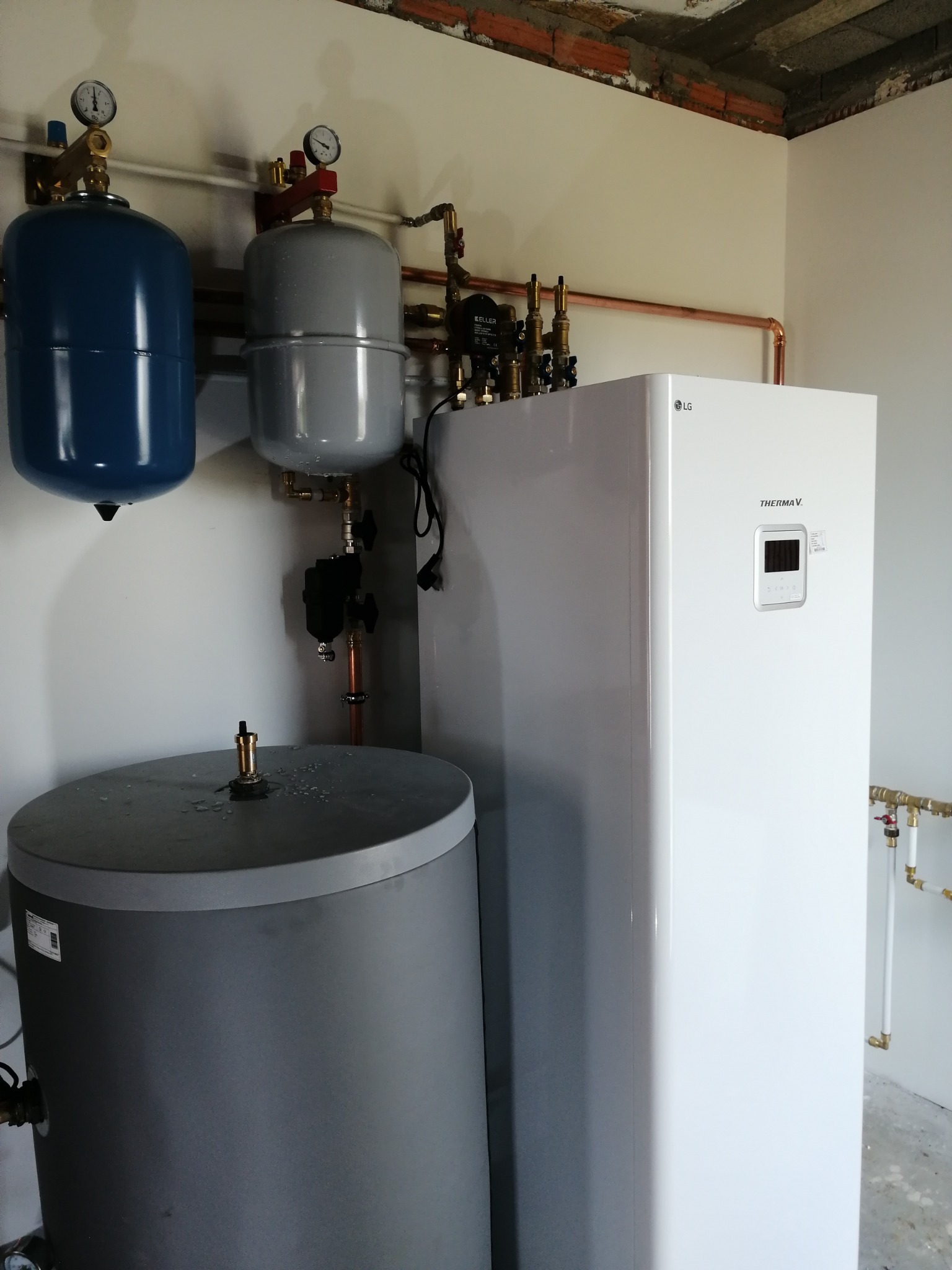 Biała pompa ciepła LG Therma V, szary zbiornik, niebieski i szary zbiornik wyrównawczy z manometrami. Miedziane rury instalacji grzewczej na ścianie.