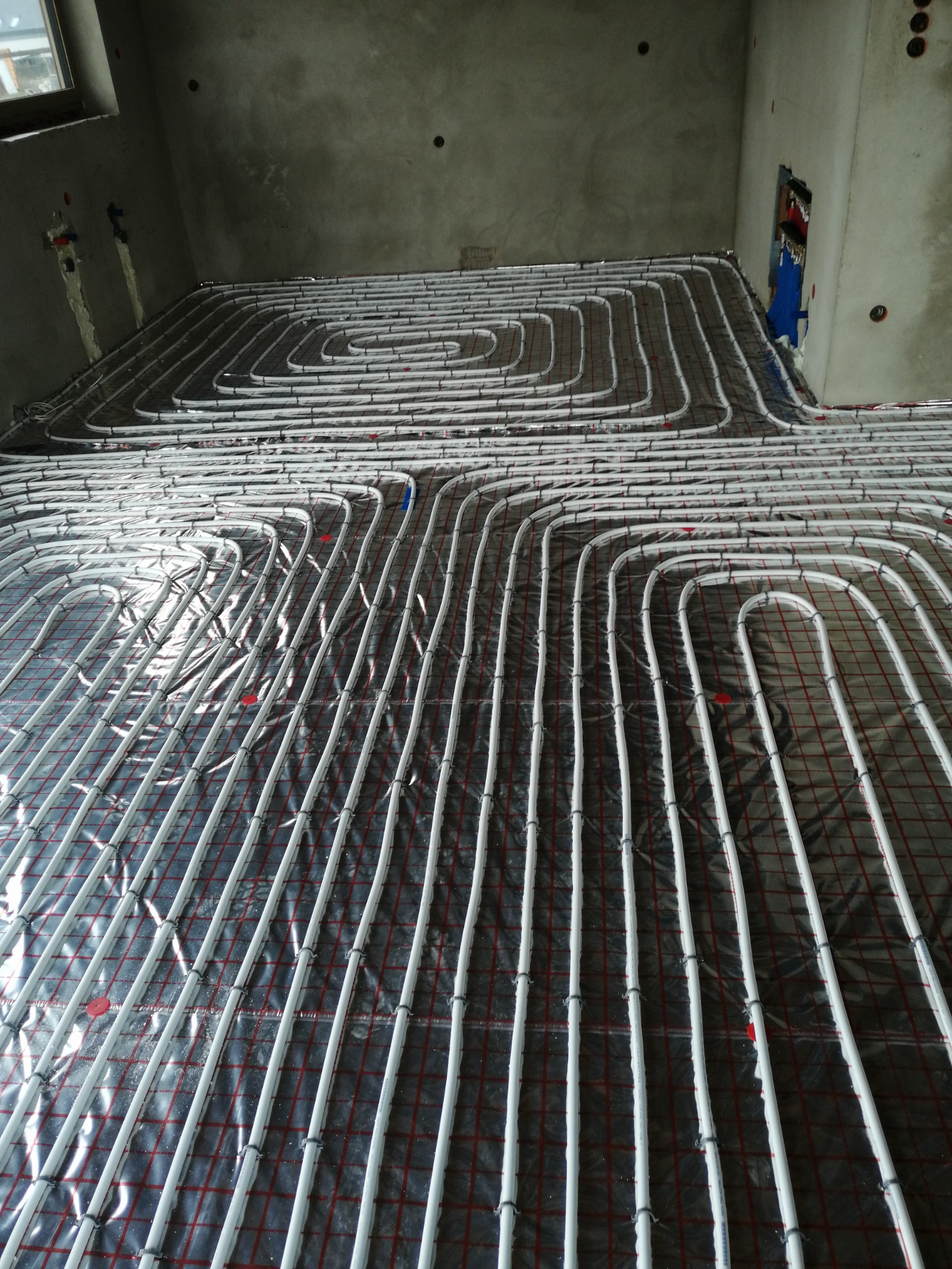 Białe rury ogrzewania podłogowego ułożone na folii z czerwoną kratą w surowym wnętrzu. Widoczne mocowania rur i fragment instalacji na ścianie.